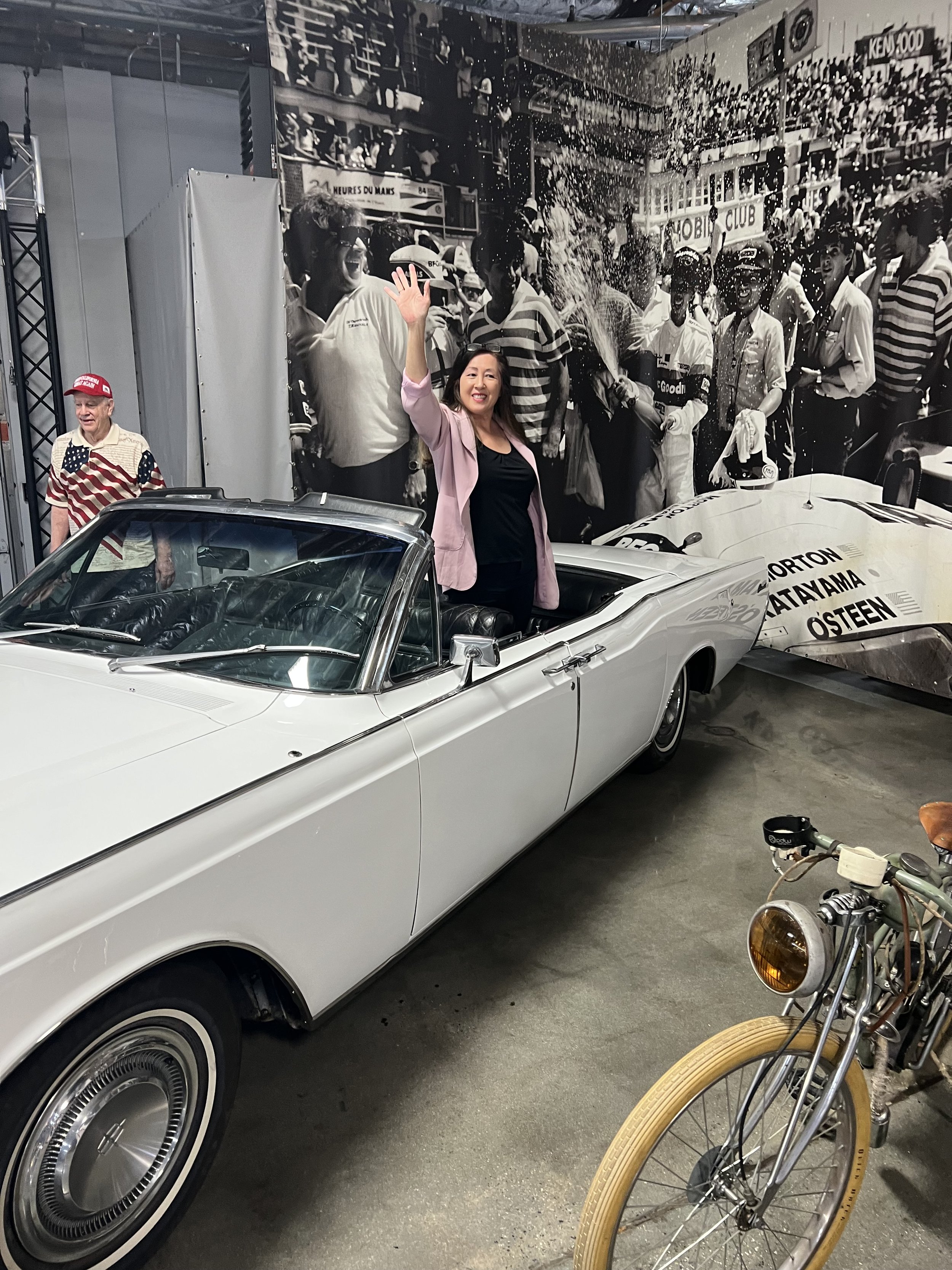 A woman in a pink blazer waving and smiling in front of a vintage white convertible car, with a large black and white mural of people celebrating and racing cars in the background. There is also a bicycle with tan tires parked nearby.