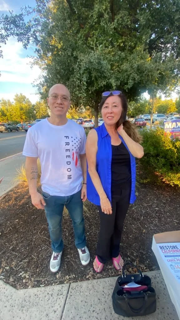A man and a woman standing outdoors near trees and parked cars, smiling at the camera on a sunny day. The man is wearing glasses, a white T-shirt with a patriotic design and jeans, while the woman is wearing a blue sleeveless shirt, black pants, and 