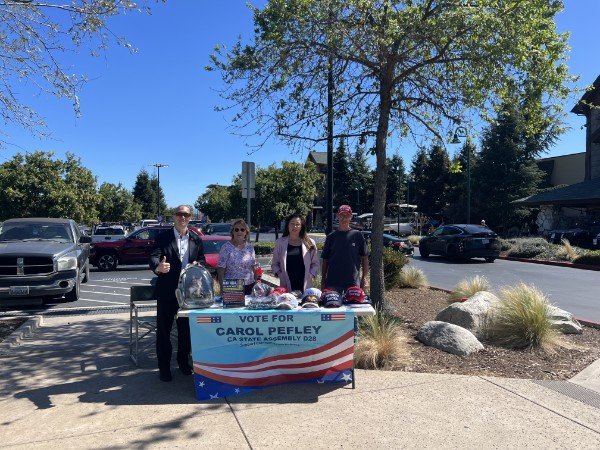 Four people standing next to a table with a sign that says "Vote for Carol Peffley, CA State Assembly D28" outside a shopping center with parked cars and trees.