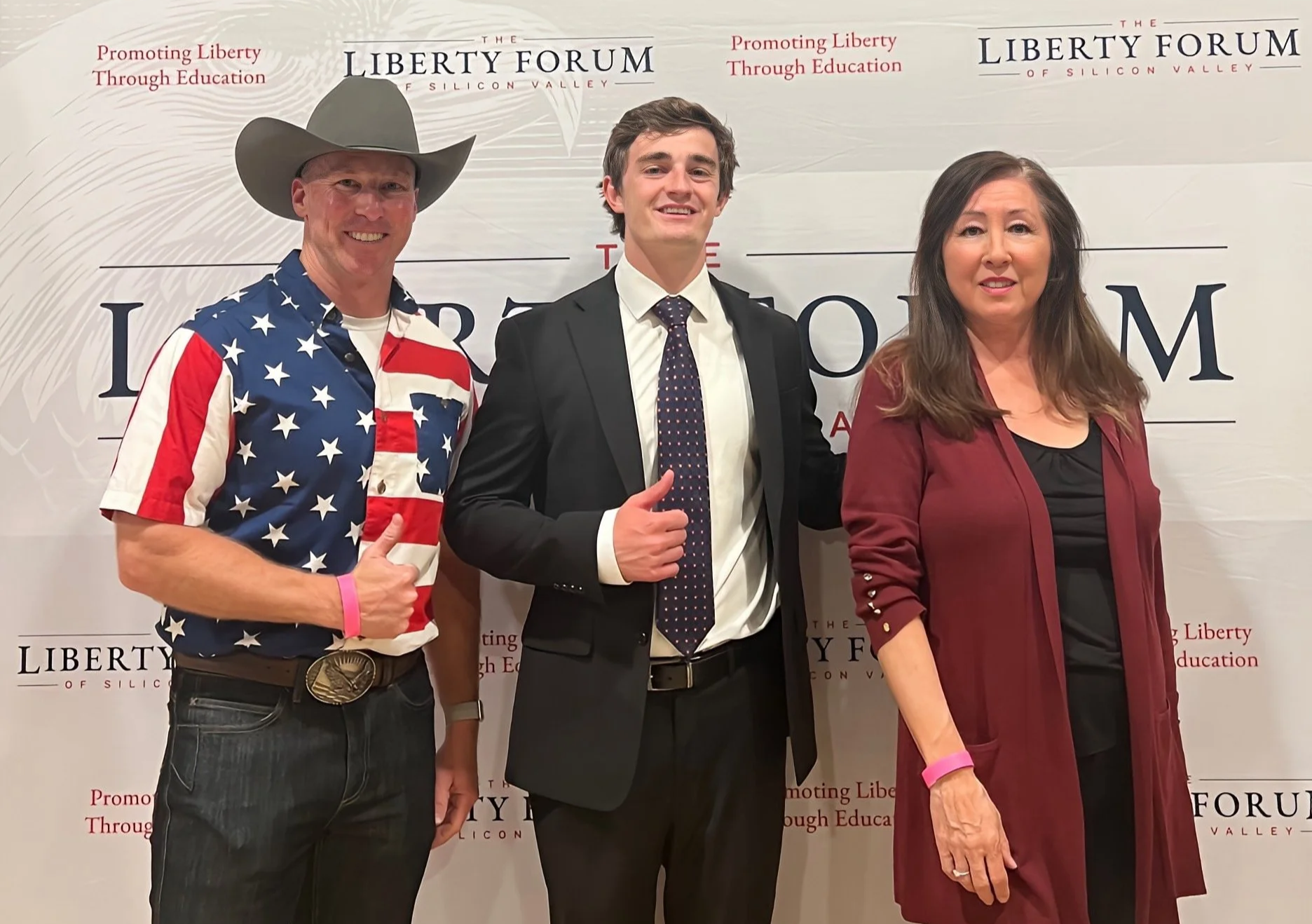 Three people standing in front of a backdrop with 'Liberty Forum of Silicon Valley' and 'Promoting Liberty Through Education' printed on it. The man on the left is wearing a cowboy hat and a shirt with an American flag pattern, the man in the middle 