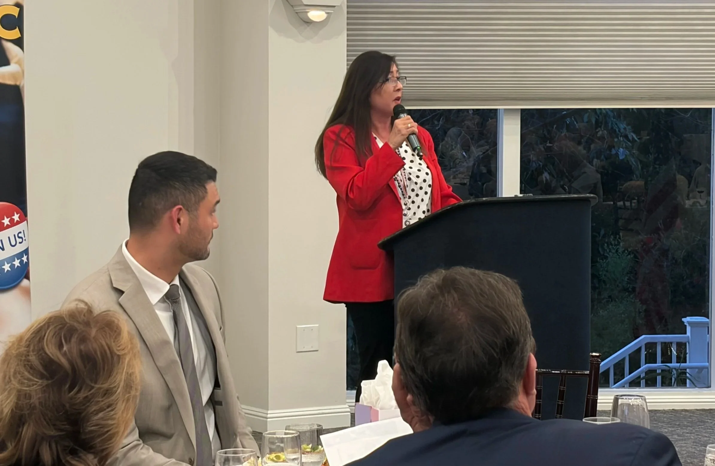A woman wearing glasses, a red blazer, and polka dot blouse is speaking into a microphone at a podium during a gathering or event. Several people are seated, listening, and some have drinks on the table in front of them. The background shows a window