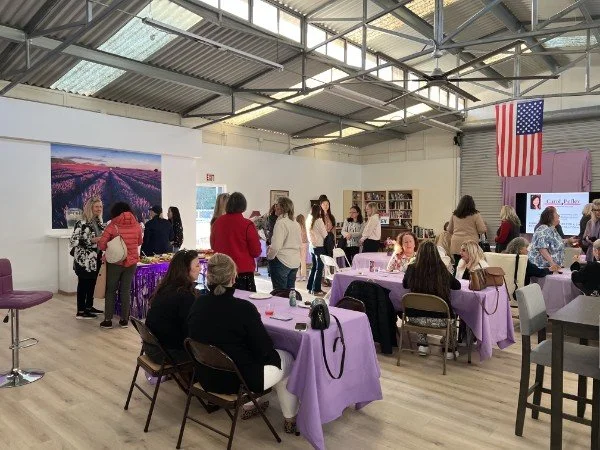 People gathered in a spacious room with purple tablecloths, some seated and some standing, attending an event, with an American flag hanging on the wall and a large landscape photograph of mountains and lavender fields.