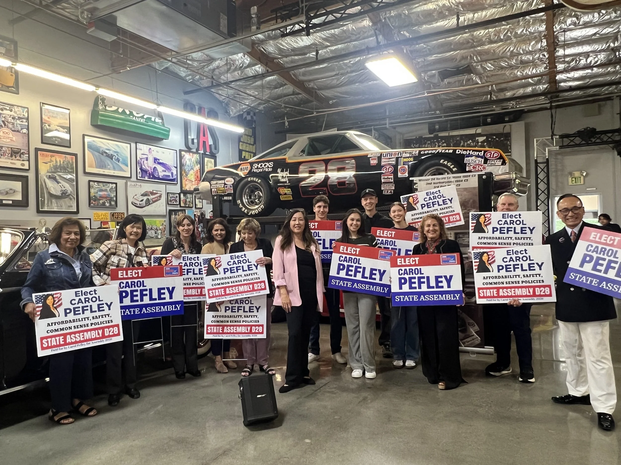 A group of people holding campaign signs supporting Carol Pefley for state assembly, standing indoors at an event with automotive posters and memorabilia on the walls, and a racing car overhead.