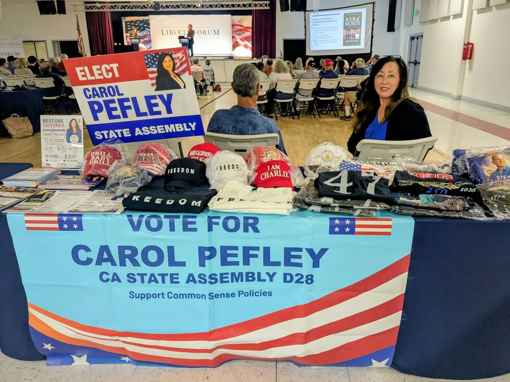 A political campaign booth supporting Carol Pefley for California State Assembly District 28. The booth features campaign signs, hats, and shirts with patriotic themes, set up inside a large auditorium with an audience watching a speaker on stage.