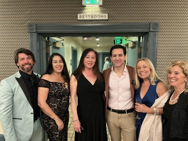 Six people standing in front of a restroom hall, smiling at the camera, with an 'EXIT' sign and a 'RESTROOMS' sign above the doorway.