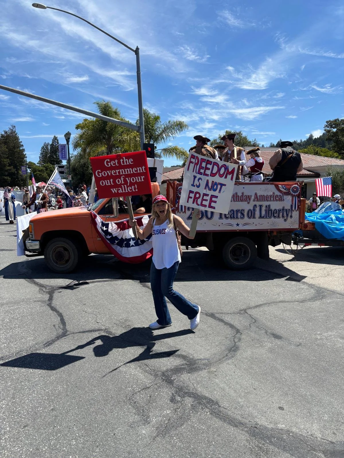 Young girl participating in a parade, holding signs that read, 'Get the Government out of your wallet!' and 'FREEDOM IS NOT FREE.' She wears a red hat and a patriotic-themed shirt, with other parade floats and people dressed in historical costumes vi