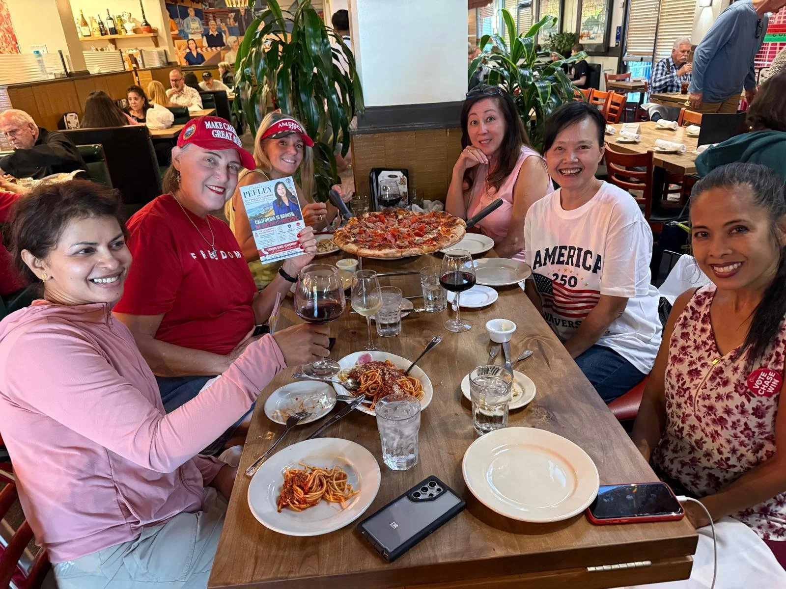 Group of women celebrating at a restaurant with pizza, pasta, and glasses of red wine, some wearing patriotic and political shirts, and campaign hats.