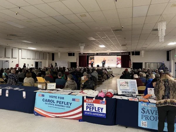 A large indoor event with many attendees seated and a speaker on stage. The room has a high ceiling with chandeliers, and there are campaign signs on a table in the foreground supporting Carol Peasley for CA State Assembly D20. A Christmas tree is vi