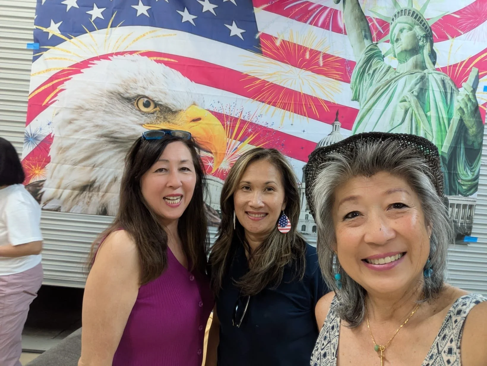 Three women smiling in front of a patriotic backdrop featuring an American flag, a bald eagle, the Statue of Liberty, and fireworks.