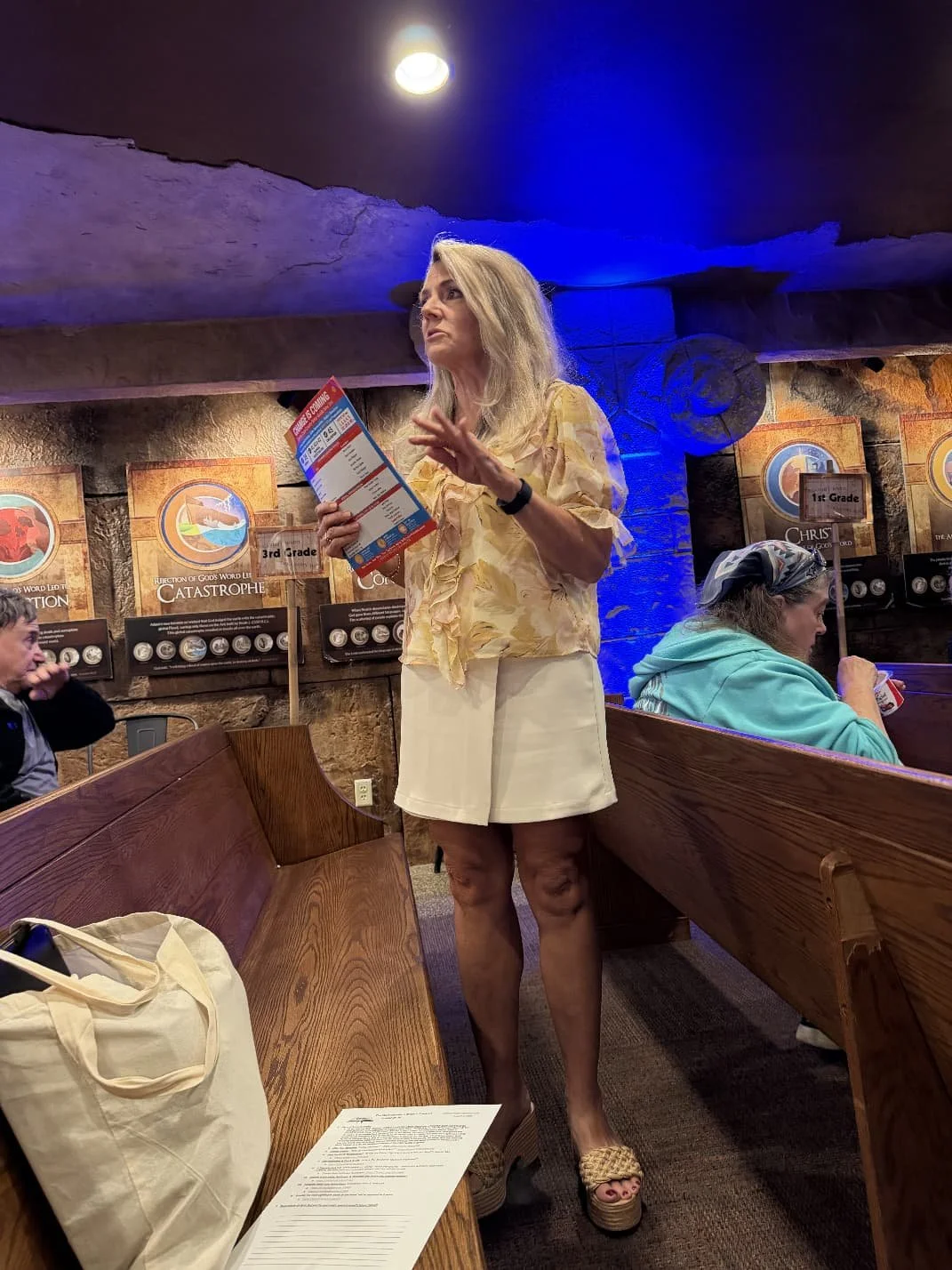 A blonde woman standing in a church or auditorium holding a brochure, wearing a light-colored floral blouse and beige skirt. Other people are seated around her, with the background displaying religious or thematic posters on the wall.
