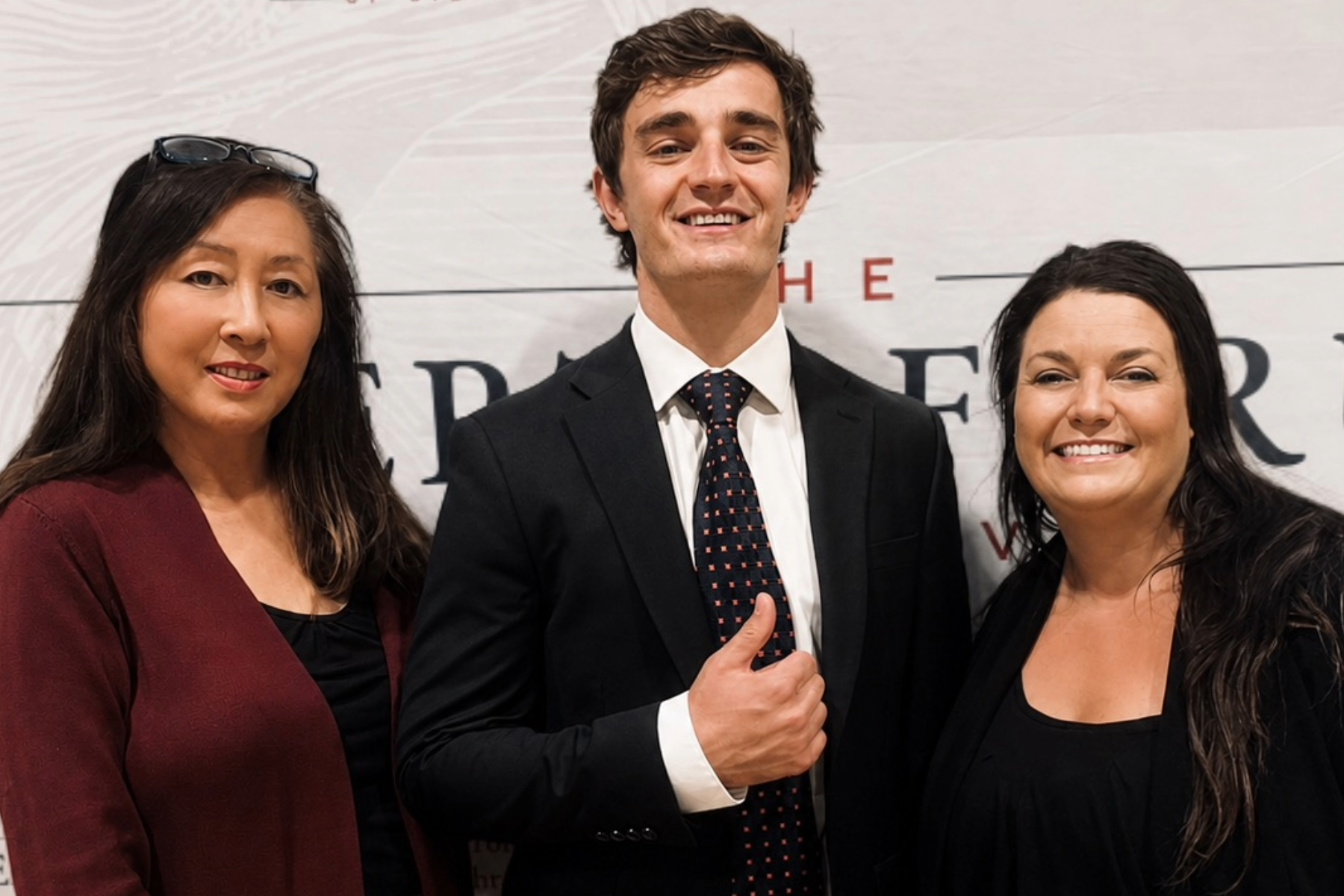 Three people posing for a photo at an event. The man in the center is wearing a black suit and tie, smiling, and giving a thumbs up. The two women on either side are smiling; the woman on the left has long dark hair, glasses on her head, and is weari