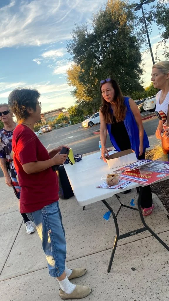 Four women gathered around a table outdoors on a sidewalk near a parking lot. One woman in a red shirt and jeans is holding items and appears to be talking to the women on the other side of the table. The table holds flyers, a box, and some cookies. 