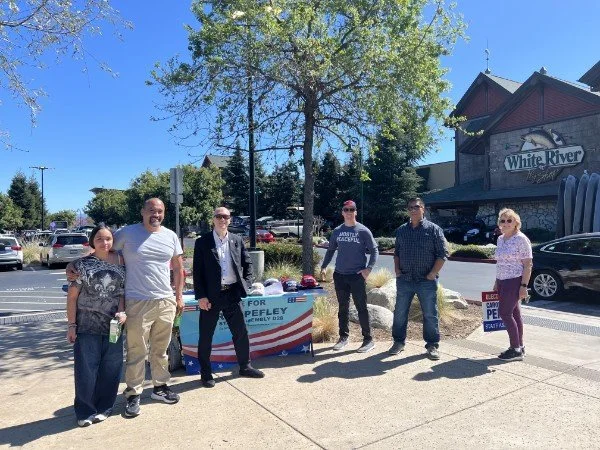 Group of six people standing outdoors next to a political campaign table with an 'Andrew Pefley' sign, in front of a building with a 'White River' sign, trees, and parking lot.