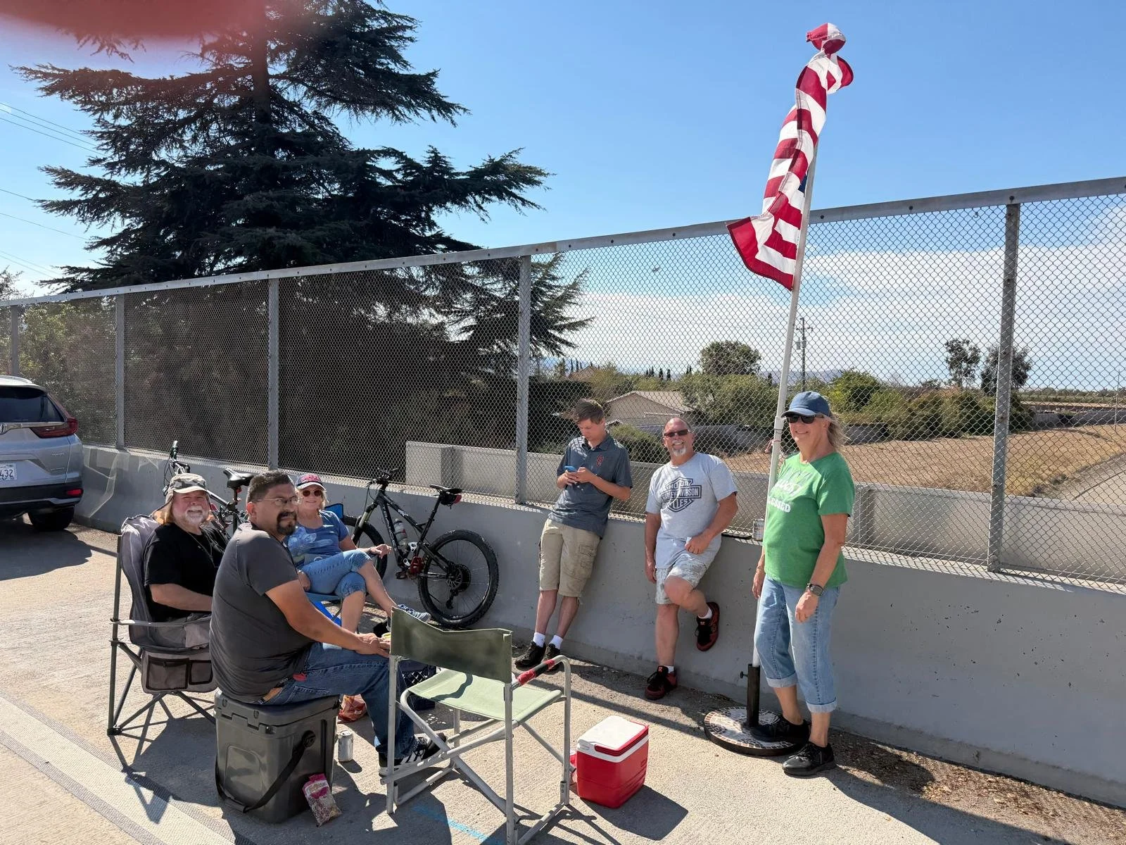 A group of six people gathered outdoors near a chain link fence with trees and houses in the background. Three are seated in folding chairs, one is standing, and two are leaning against the fence. There are bicycles and a red cooler, and a woman is h