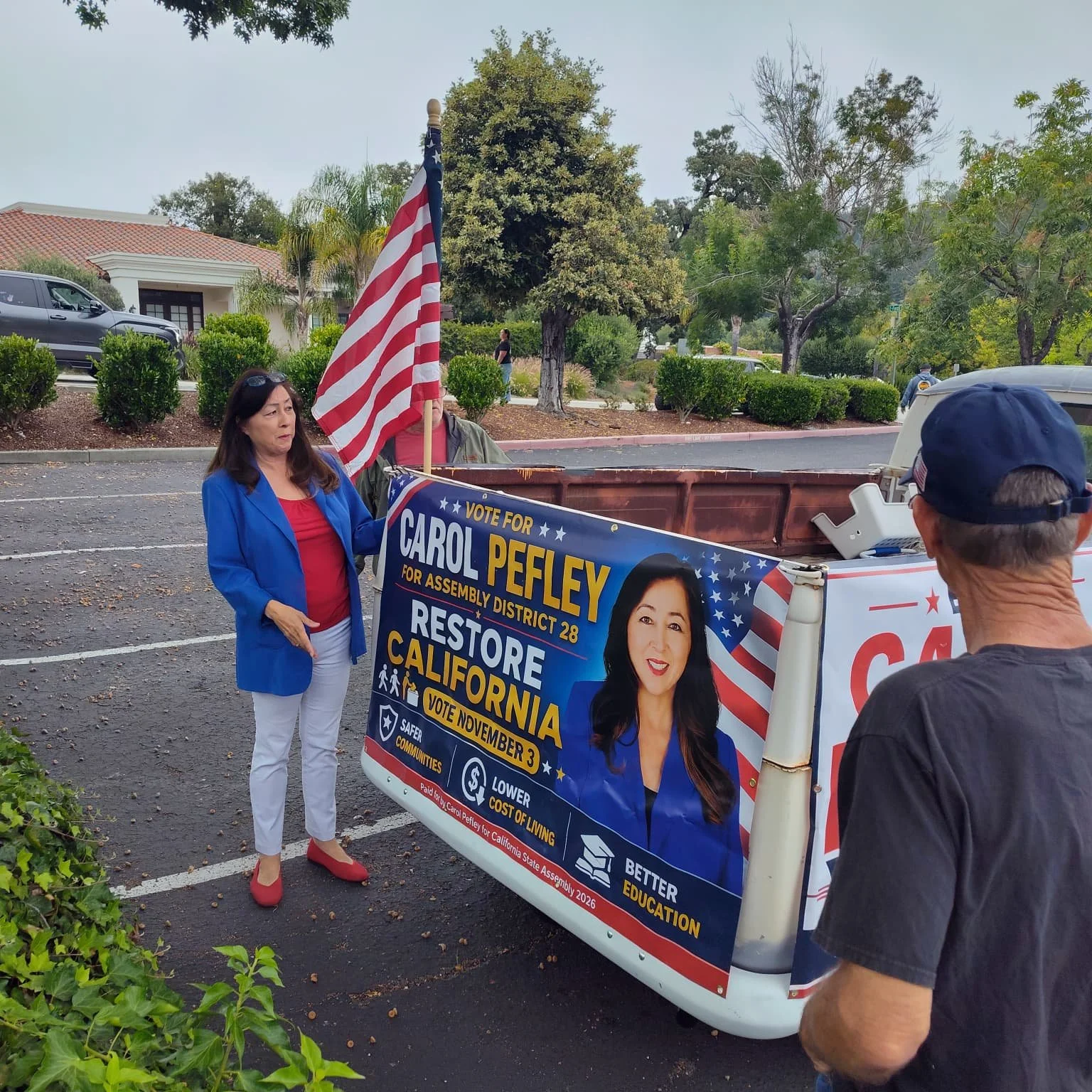 Carol Pefley in blue blazer and red shoes standing next to political campaign sign and flag, talking to man in black cap outside in a parking lot with trees and buildings in the background.