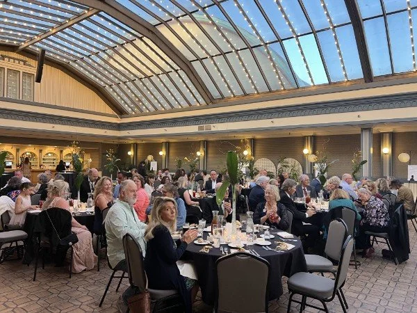 People dining at round tables in a banquet hall with a glass ceiling and decorated walls.