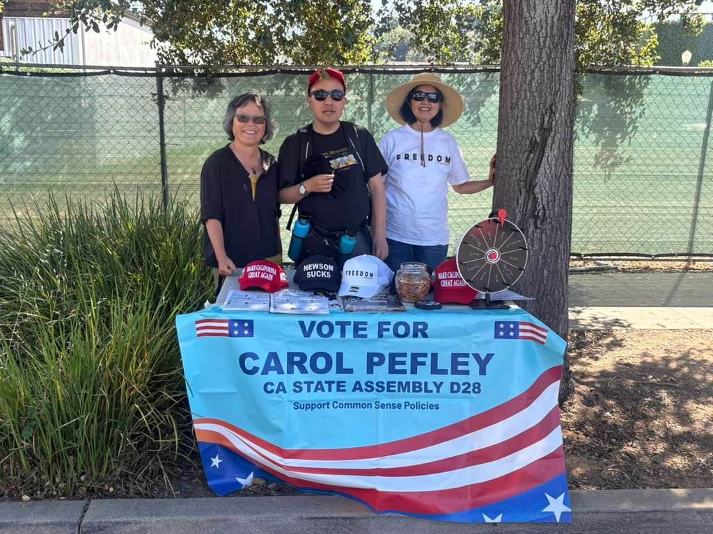 Three people standing behind a table with campaign materials supporting Carol Pefley for CA State Assembly D28. The table has a blue banner with red and white stripes, stars, and the text 'VOTE FOR CAROL PEFLEY CA STATE ASSEMBLY D28 Support Common Se
