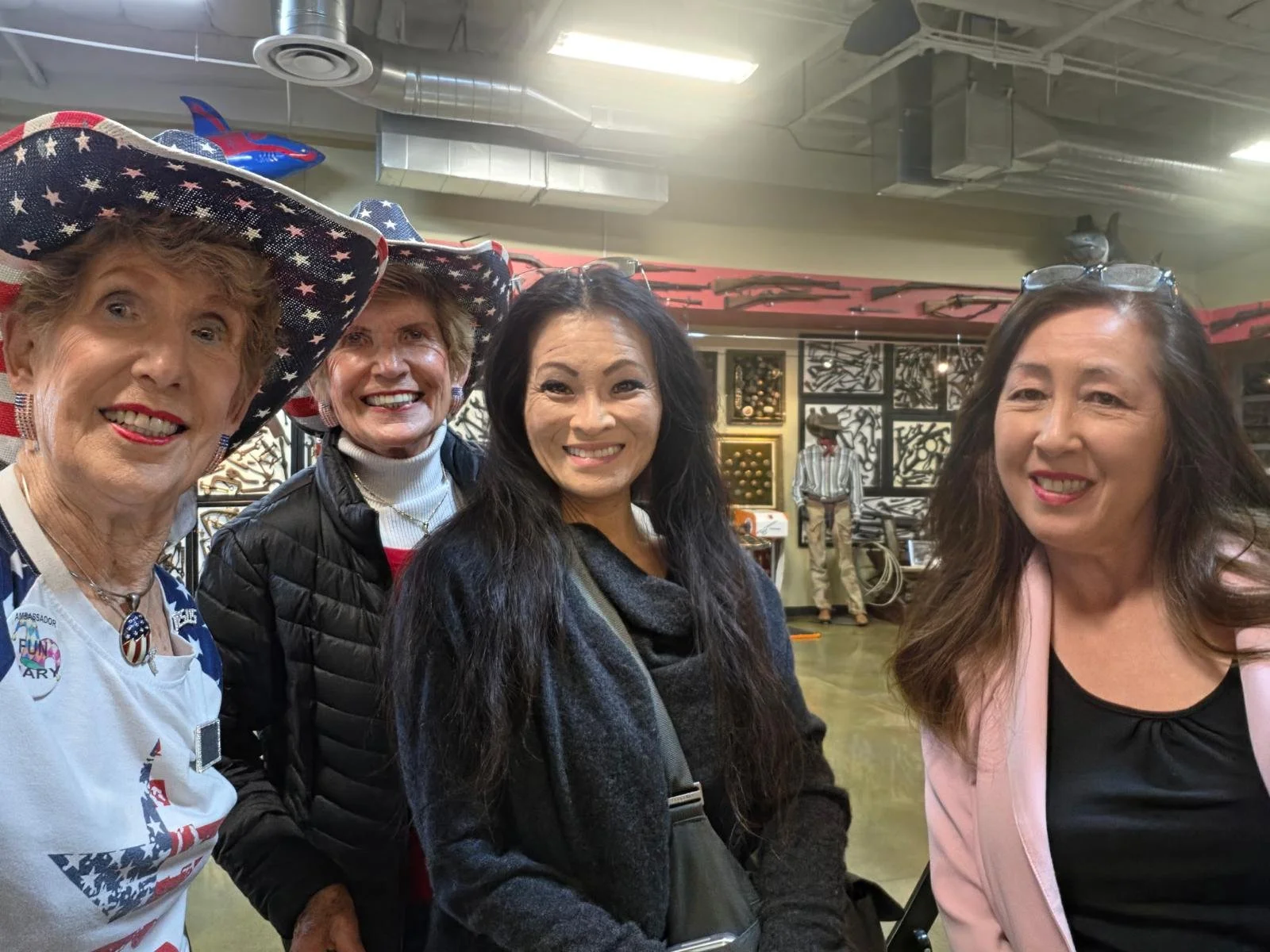 Four women smiling at the camera inside a room decorated with patriotic and military-themed items, including rifles and artwork on the walls.
