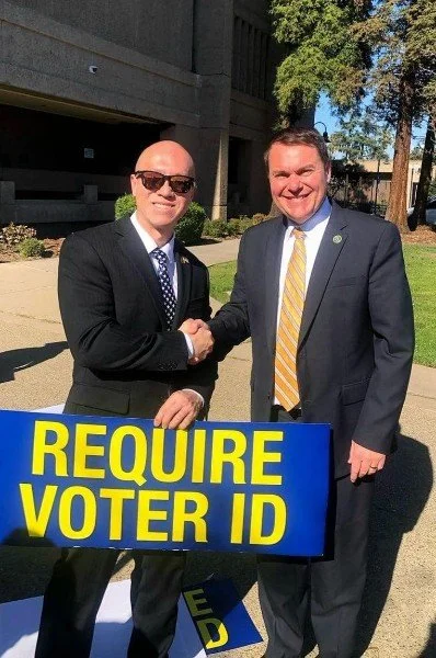 Two men in suits shake hands outdoors near a sign that reads "REQUIRE VOTER ID."