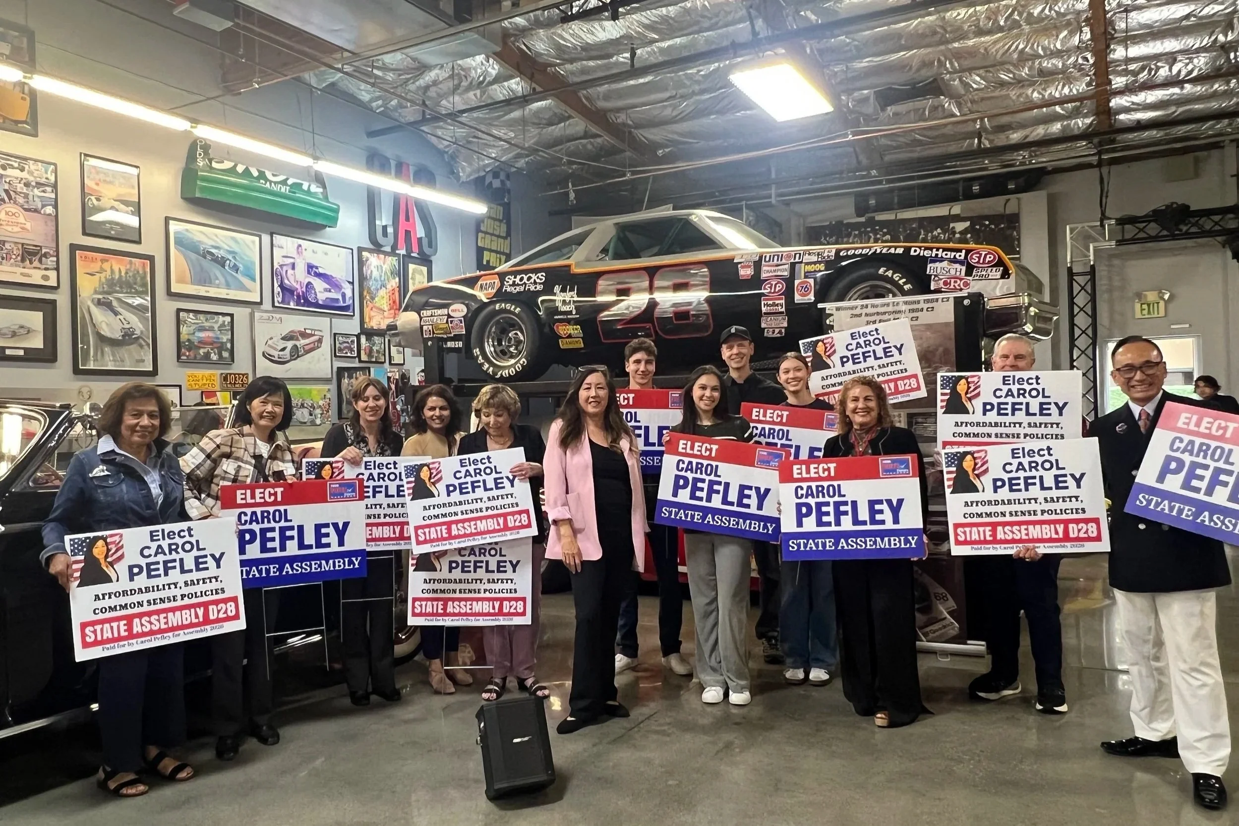 Group of people holding signs supporting election candidate Carol Pefley in an indoor car museum. The signs promote affordability, safety, and common sense policies for State Assembly D28. The background features car artwork and a race car displayed on the wall.