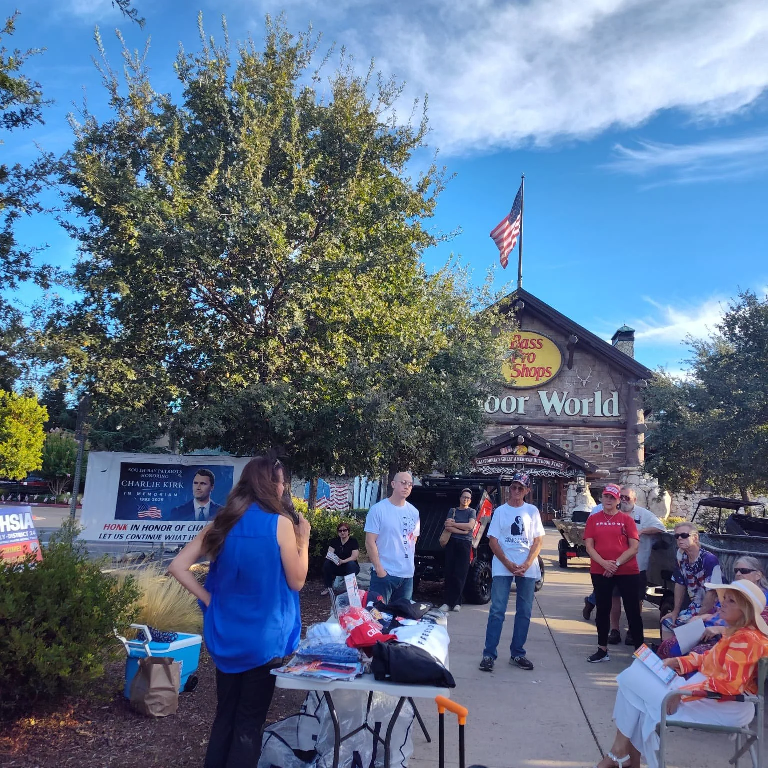A group of people gathered outside the Bass Pro Shops store in outdoor clothing, with some seated and others standing, near a table with promotional materials, in front of the store's entrance which has trees, a blue sky, and American flags.
