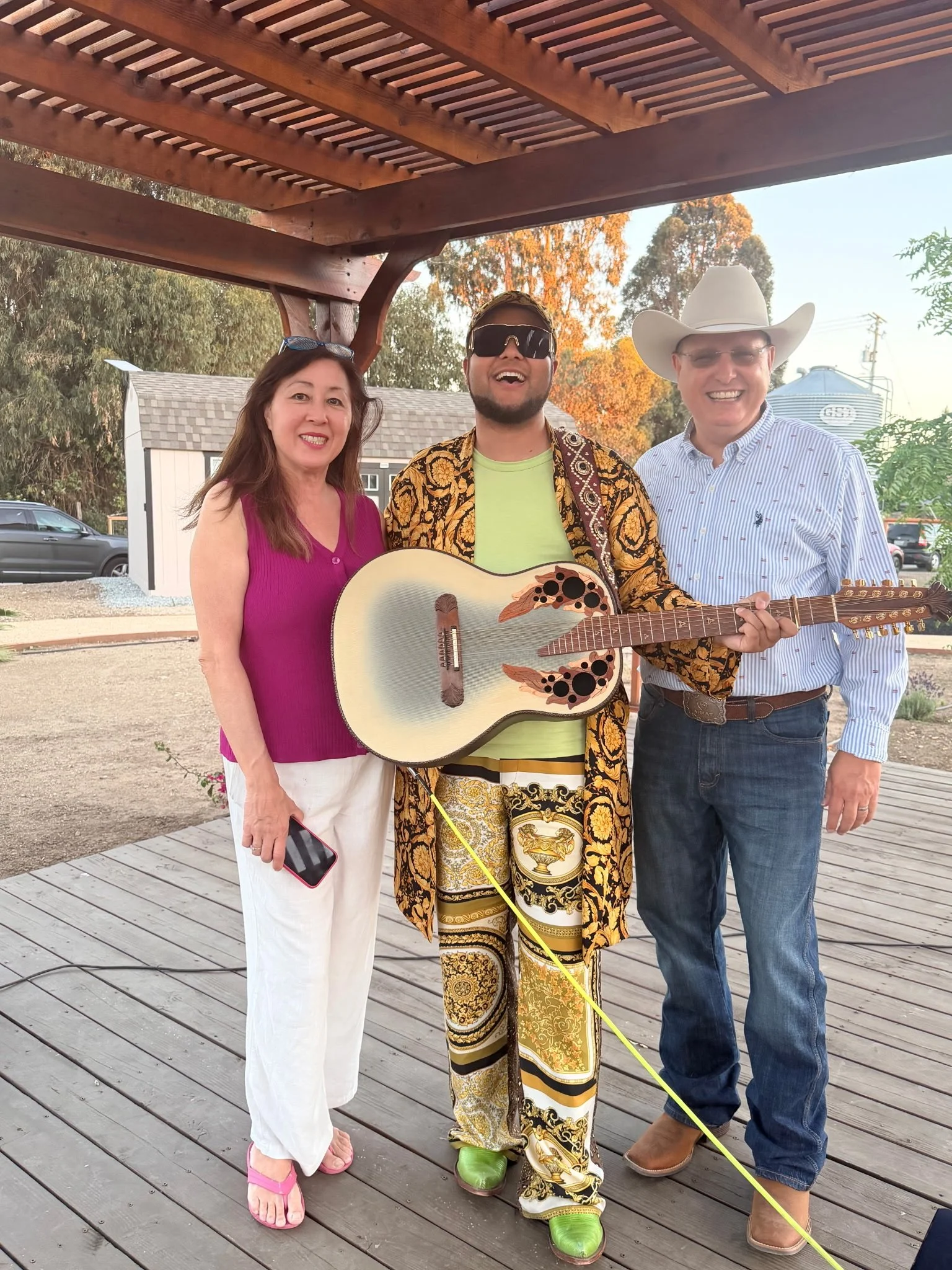 Three people standing under a wooden shelter, smiling. The person in the middle is wearing a patterned outfit, large sunglasses, and holding a guitar. The person on the right is wearing a cowboy hat, glasses, a button-up shirt, and jeans. The person 