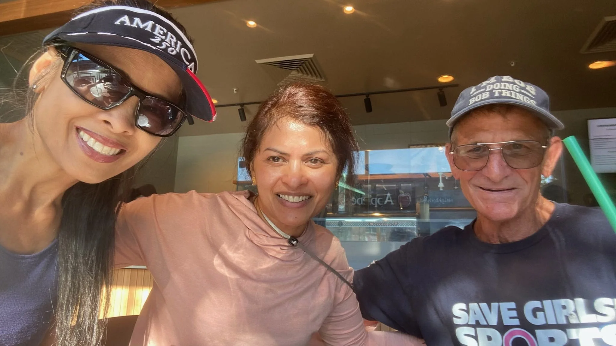 Three people smiling for a selfie at a restaurant or cafe. The woman on the left is wearing sunglasses and a headband, the woman in the middle has short brown hair, and the man on the right is wearing glasses, a baseball cap, and a shirt with visible