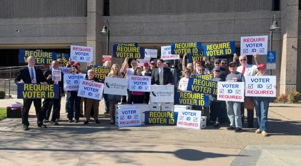 Group of people gathered outdoors holding signs that say "Require Voter ID" and "Voter ID Required" to promote voting requirements.