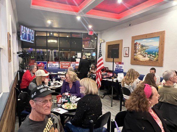 A group of people sitting at tables in a restaurant or community room with political signs and an American flag, engaged in conversation.