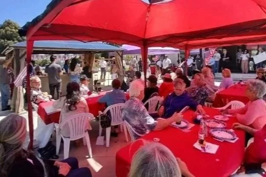 People gathered at an outdoor event, seated under red and purple tents, with tables set for a meal, some holding drinks, and American flags displayed in the background.