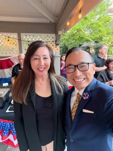 Two people taking a selfie at an outdoor event with group of people in the background, decorated with American flags and string lights.