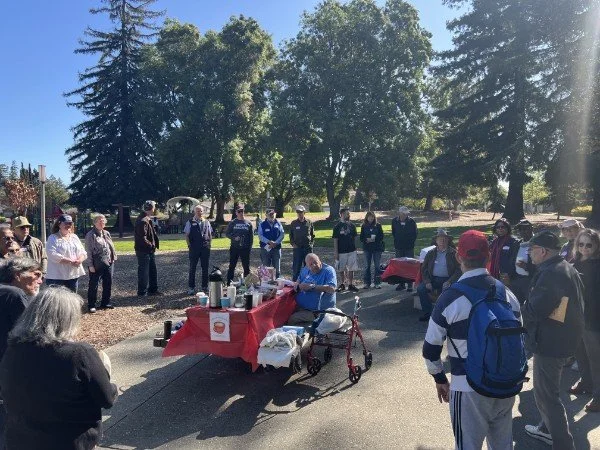 Group of people gathered outdoors around a table with food and drinks, under trees on a sunny day, with one person sitting with a walker.