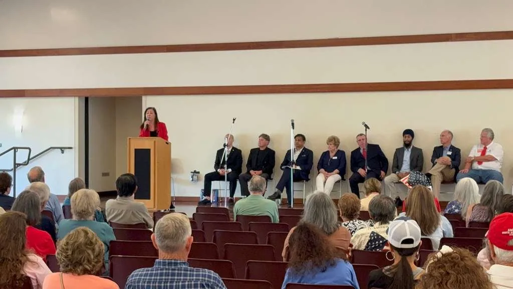 A panel discussion with a woman speaking at a podium on stage, seven people seated behind her, and an audience in a conference room.
