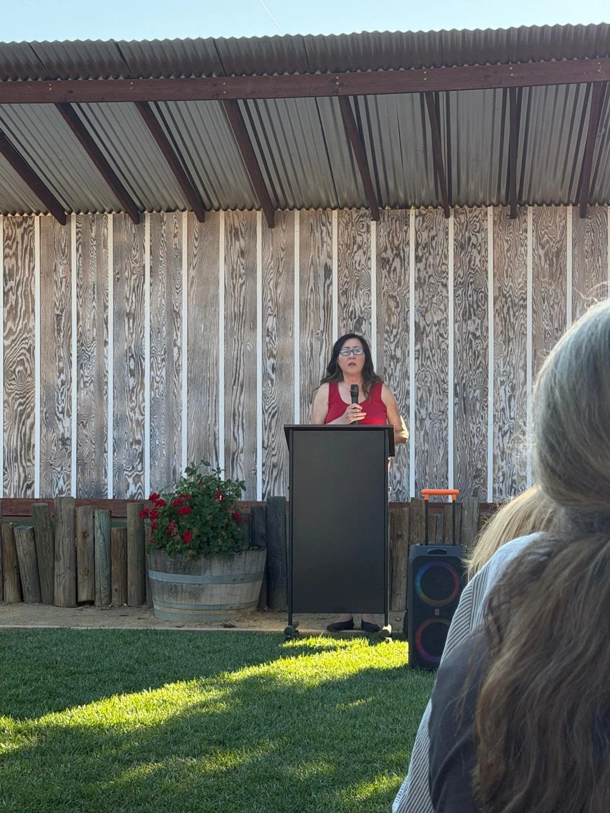 A woman in a red dress with glasses speaking into a microphone at an outdoor event, standing behind a black podium. There are people seated in front, a wooden fence, and a wooden barrel with red flowers. The background features a wooden wall and a co