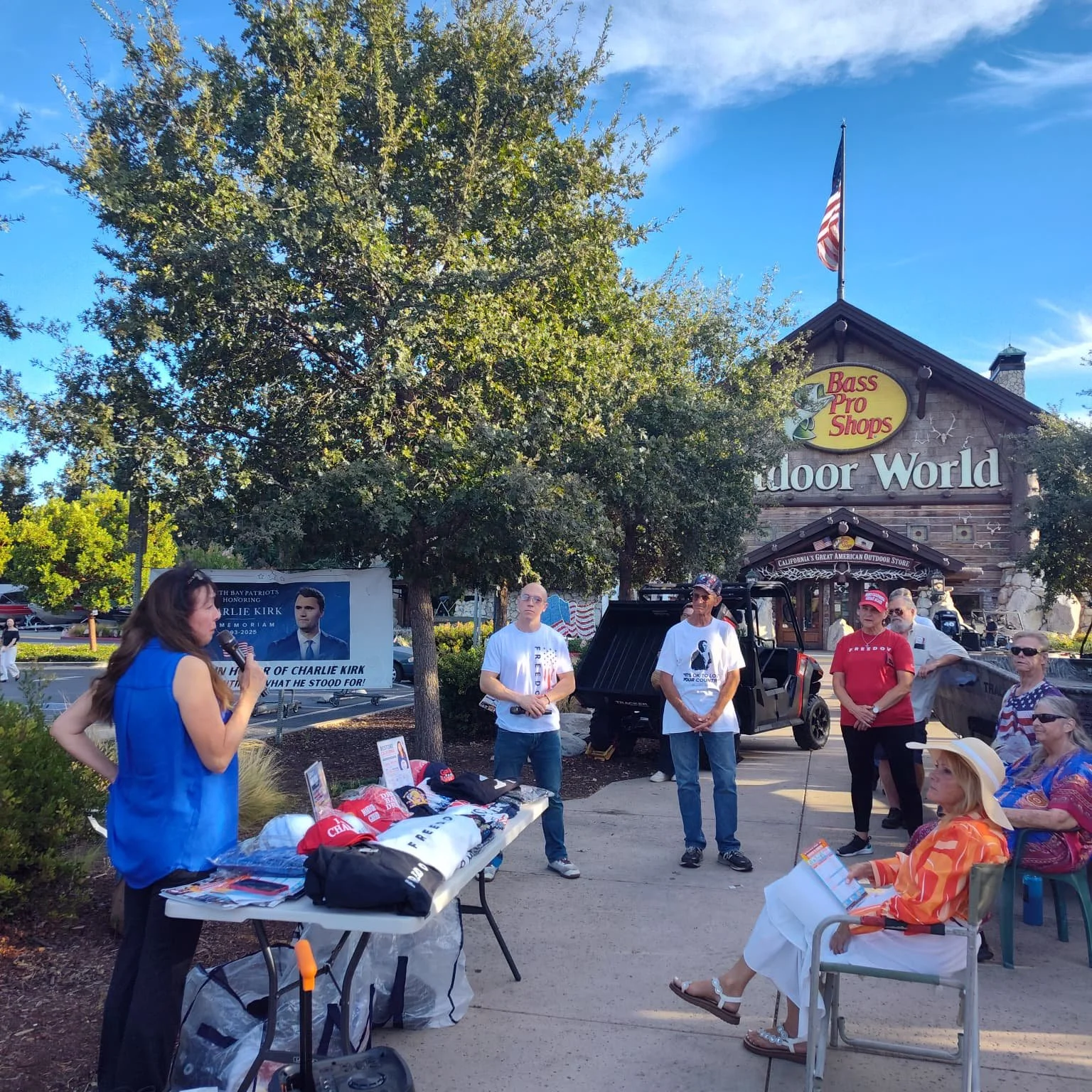 A group of people gathered outdoors under a tree in front of a Bass Pro Shops store, participating in what appears to be a meeting or event. There is a woman speaking into a microphone, several individuals listening, and a table with merchandise and 