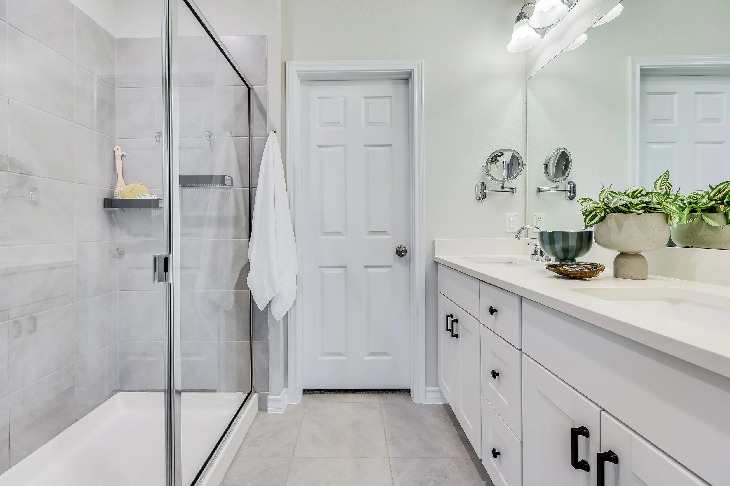 5216 Golden Canary Lane Austin, TX 78723. Bathroom with a glass shower door, a white vanity with black hardware, a large mirror, two small round magnifying mirrors on the wall, a potted plant, decorative bowls, and a closed white door.