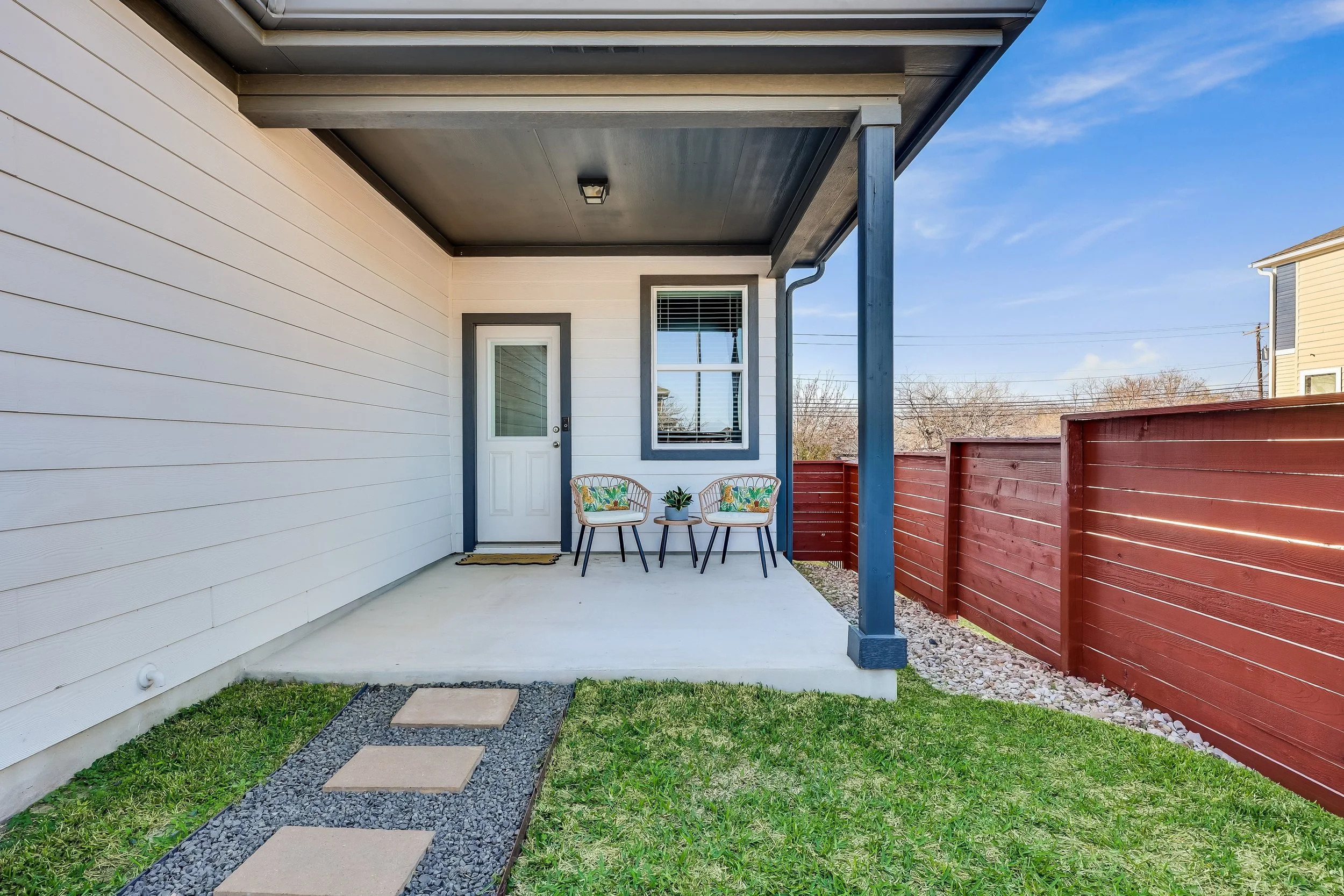 5216 Golden Canary Lane Austin, TX 78723. Front porch of a house with white siding, a small seating area with two chairs and a side table, a concrete slab, a red wooden privacy fence, and a grass yard under a blue sky.