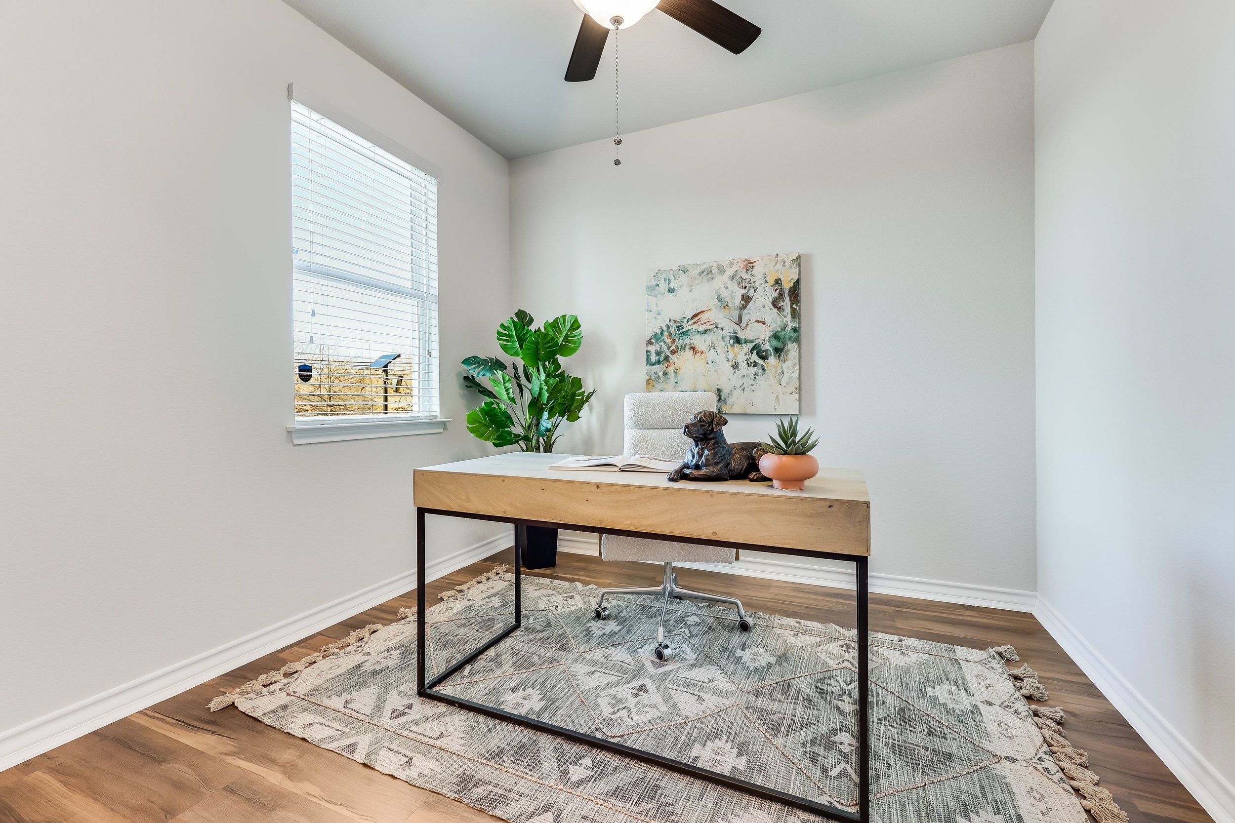 5216 Golden Canary Lane Austin, TX 78723. A modern office room with a wooden desk, a white office chair, a potted plant, a sculpture of a dog, a small potted succulent, and a large abstract painting on the wall. There's a window with white blinds. 