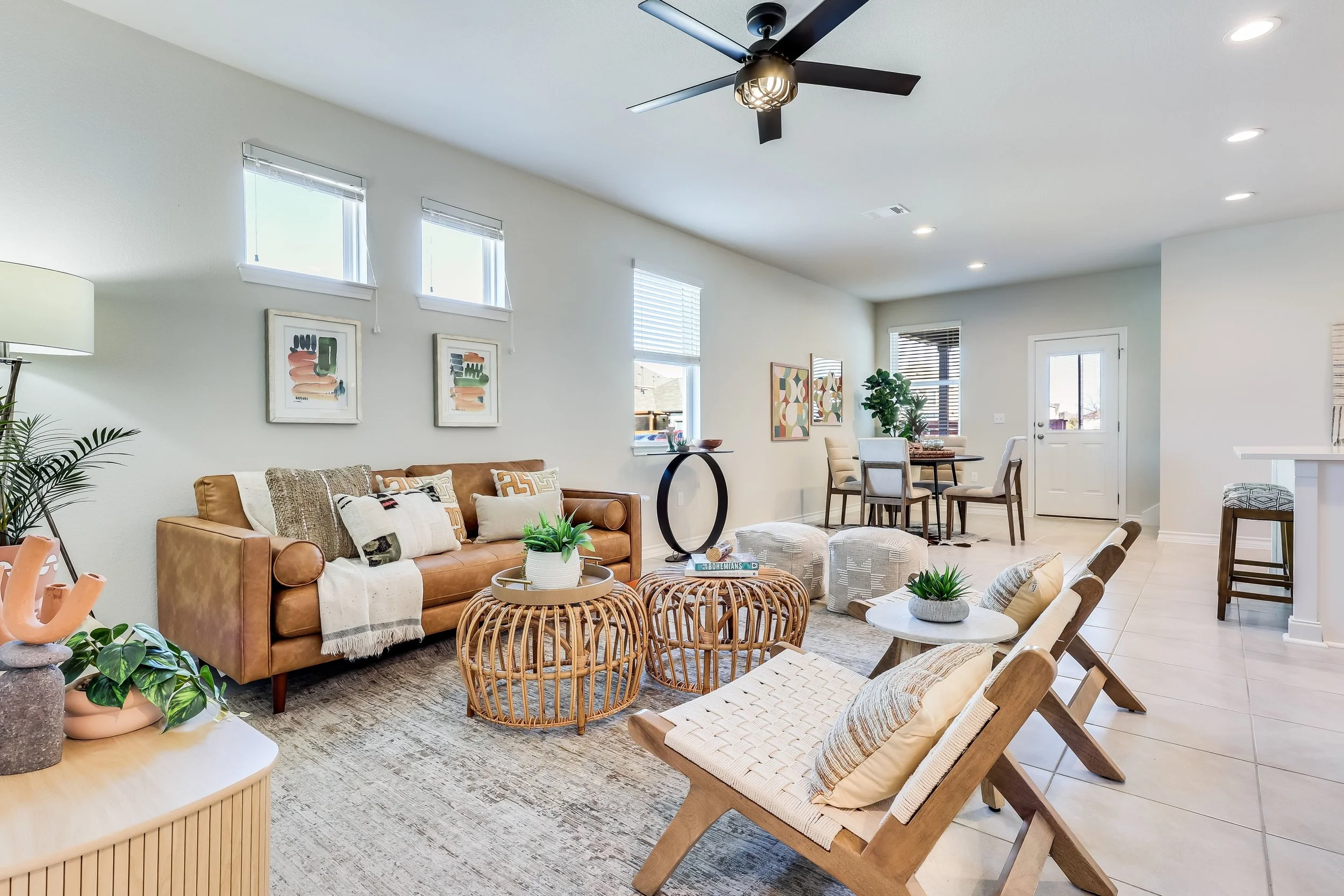 5216 Golden Canary Lane Austin. A modern living room with a brown leather sofa, beige chairs, and a round coffee table, decorated with plants, abstract art, and cushions. Open space with white walls, ceiling fan, and a dining area in the background.