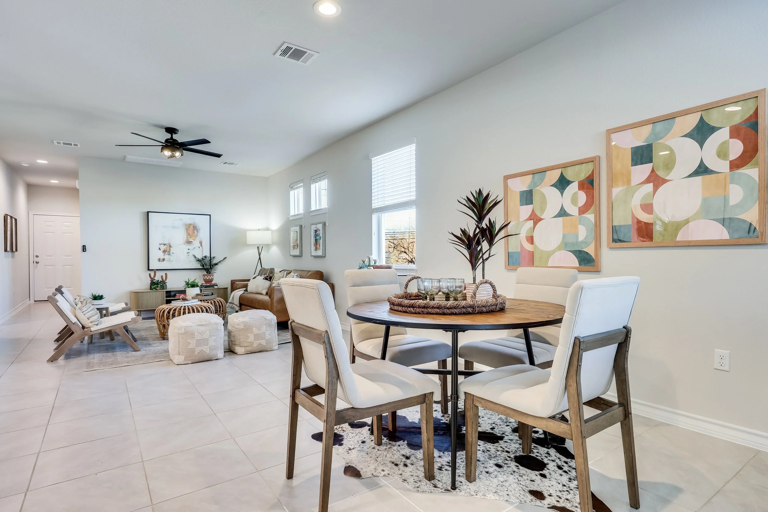 Open concept living and dining room with white walls, large windows, and modern furniture. The dining table has four white cushioned chairs, and the living area features a leather sofa, armchair, potted plants, and wall art. There is a ceiling fan an