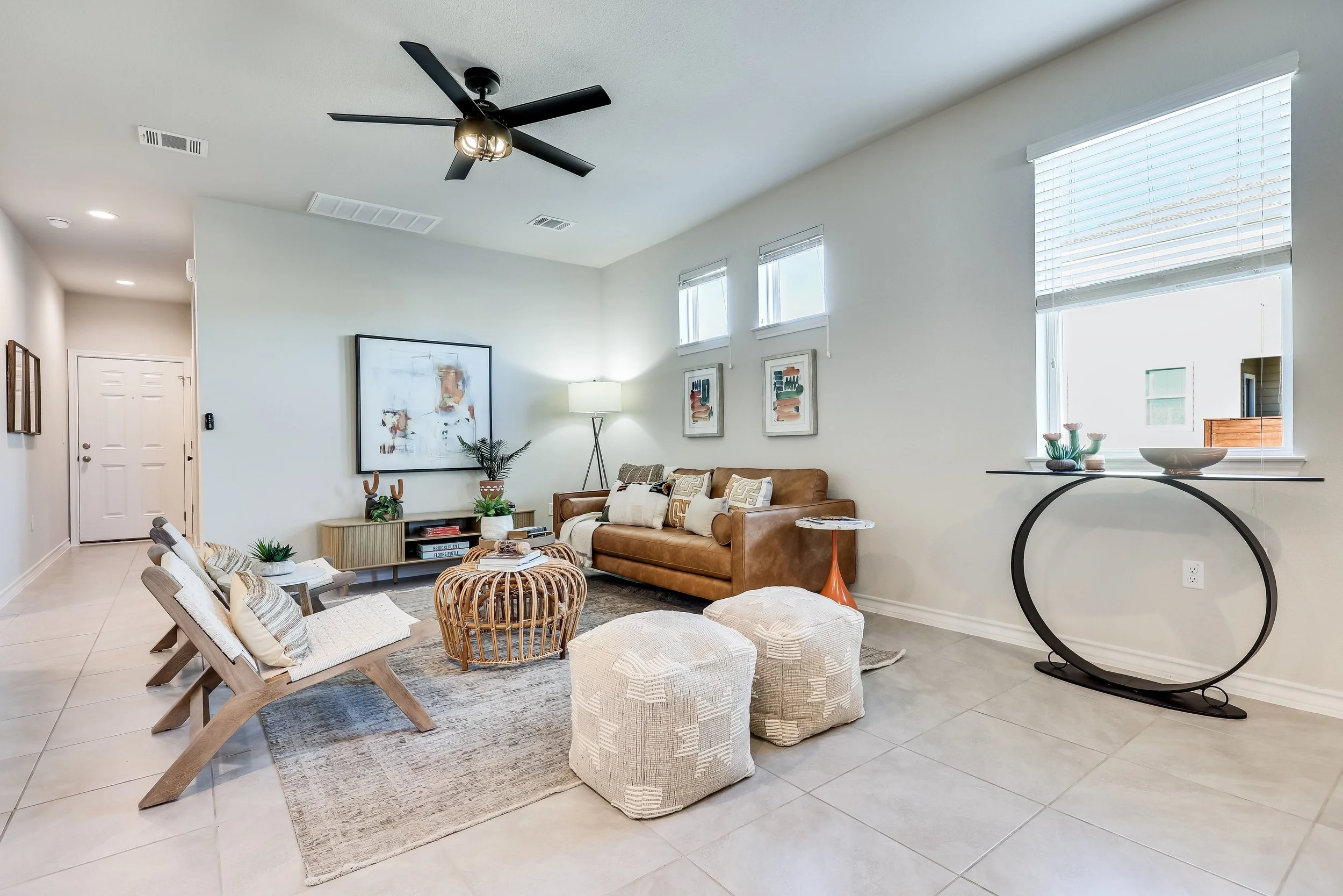 5216 Golden Canary Lane Austin, TX 78723. Living room with a brown leather sofa, wooden chairs with cushions, a round rattan coffee table, two cream ottomans, modern art on the walls, a floor lamp, and a black ceiling fan.
