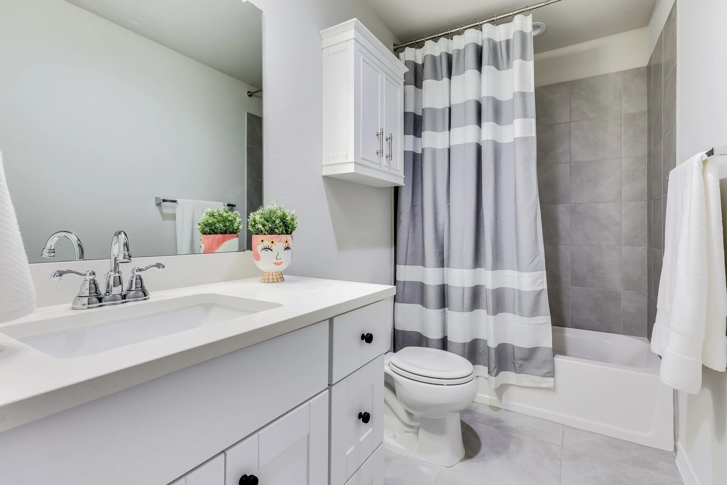 5216 Golden Canary Lane Austin, TX 78723. A modern bathroom with a white vanity, a large mirror, a sink with chrome fixtures, two potted plants with face designs on the counter, a white toilet, a striped gray and white shower curtain, a bathtub.