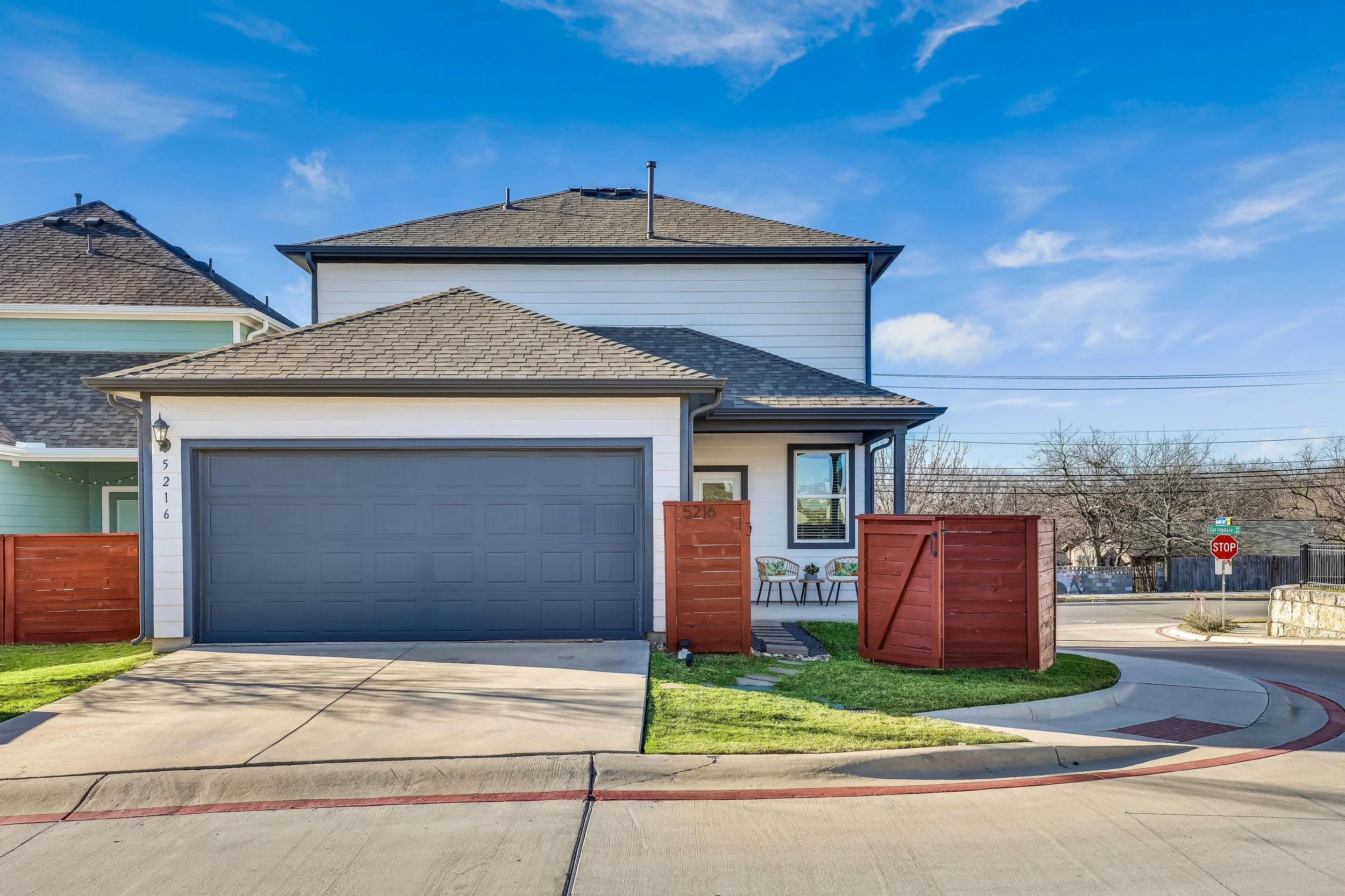 A modern two-story house with a gray garage door, white siding, and a small patio with two chairs and a plant. The house has a brown shingled roof and a red wooden fence around the small garden and entrance. The street view includes a stop sign at th