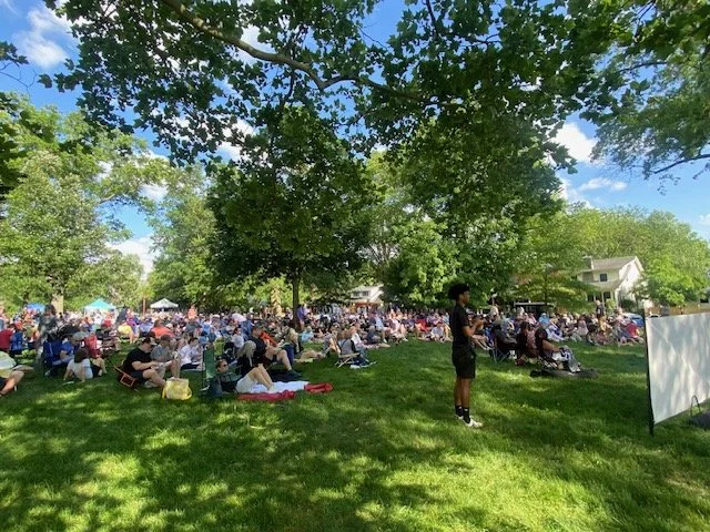 Large group of people gathered outdoors on a grassy area under trees, attending an event with many chairs and some standing people. There is a white screen on the right and houses in the background, with a clear blue sky overhead.