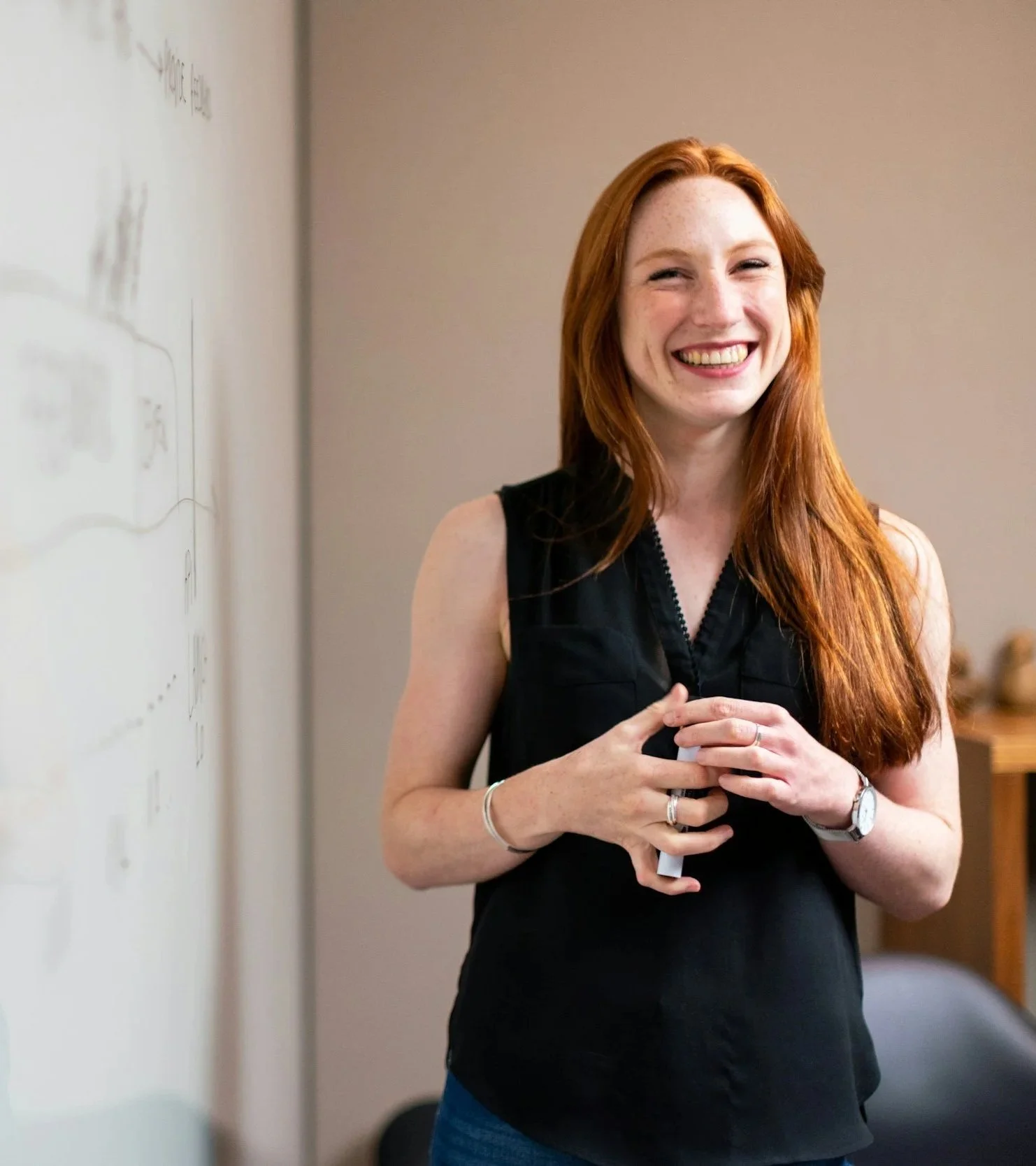 A young woman with long red hair smiling and standing in front of a whiteboard, holding a marker in her hands.