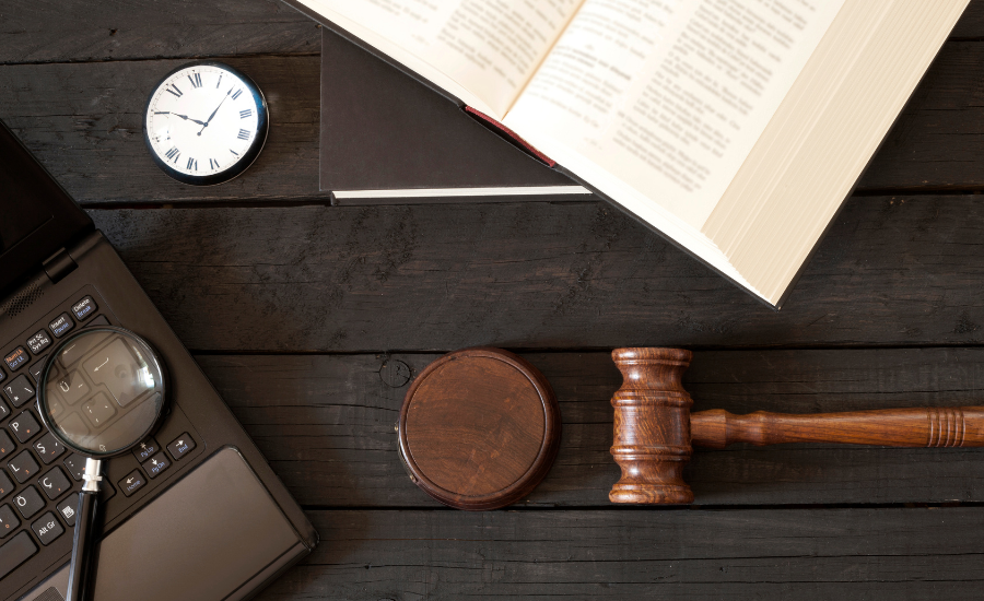 A desk with a laptop, a magnifying glass, a round wooden gavel block, a judge's gavel, an open book, a closed notebook, and a clock showing 9:09.