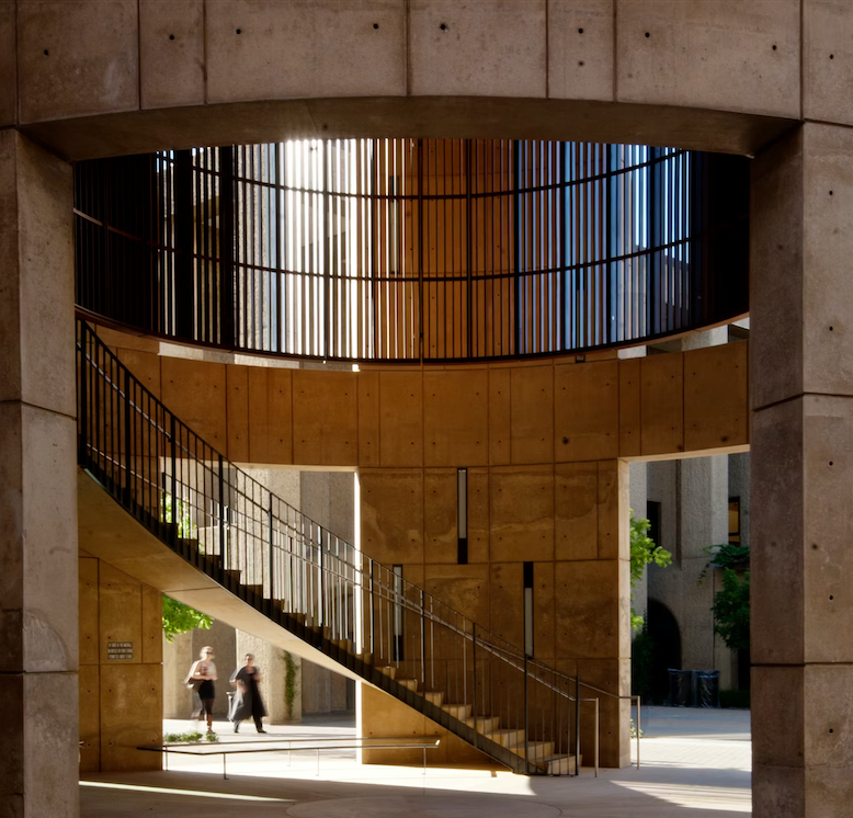 Interior view of a modern building with concrete walls, featuring a curved staircase with black railings, illuminated by sunlight from openings above and behind.