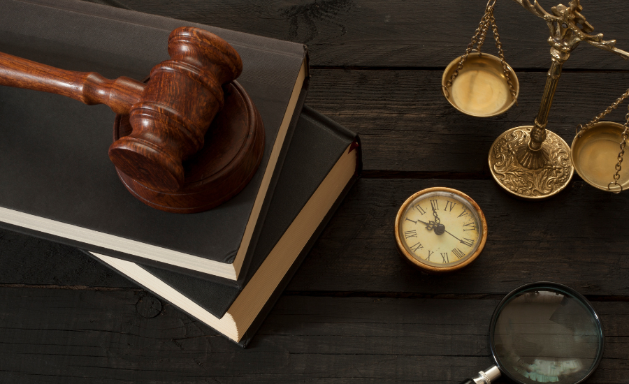 A wooden judge's gavel resting on two stacked law books, a brass balance scale, a small round clock, and a magnifying glass on a dark wooden surface.
