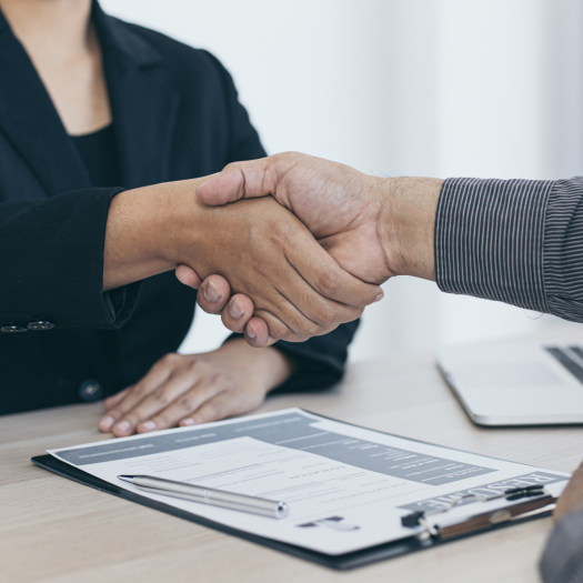 Two people shaking hands across a desk with documents and a laptop.