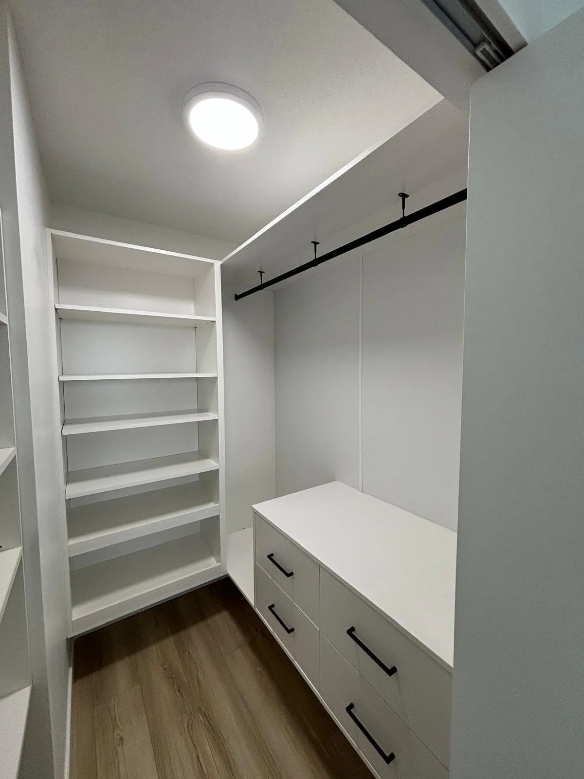 Empty walk-in closet with white shelves, drawers, and a hanging rod, with wood flooring and a ceiling light.